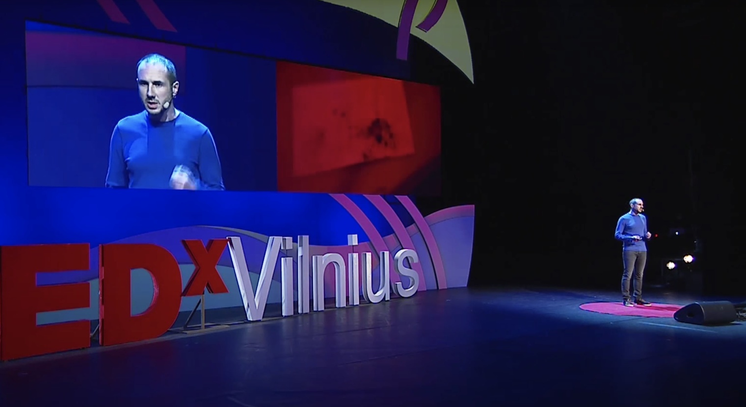 A man speaking on a large stage in front of a screen showing a realtime feed of the speech and a large red TEDx logo.